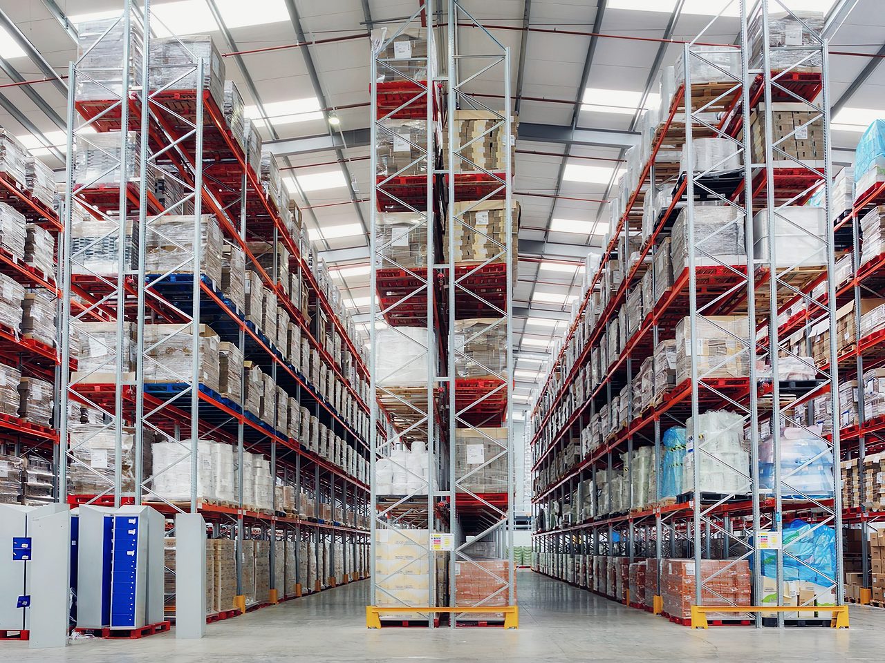 Pasillo de un depósito con estanterías selectivas de gran altura, largueros rojos y pallets cargados en todos los niveles.