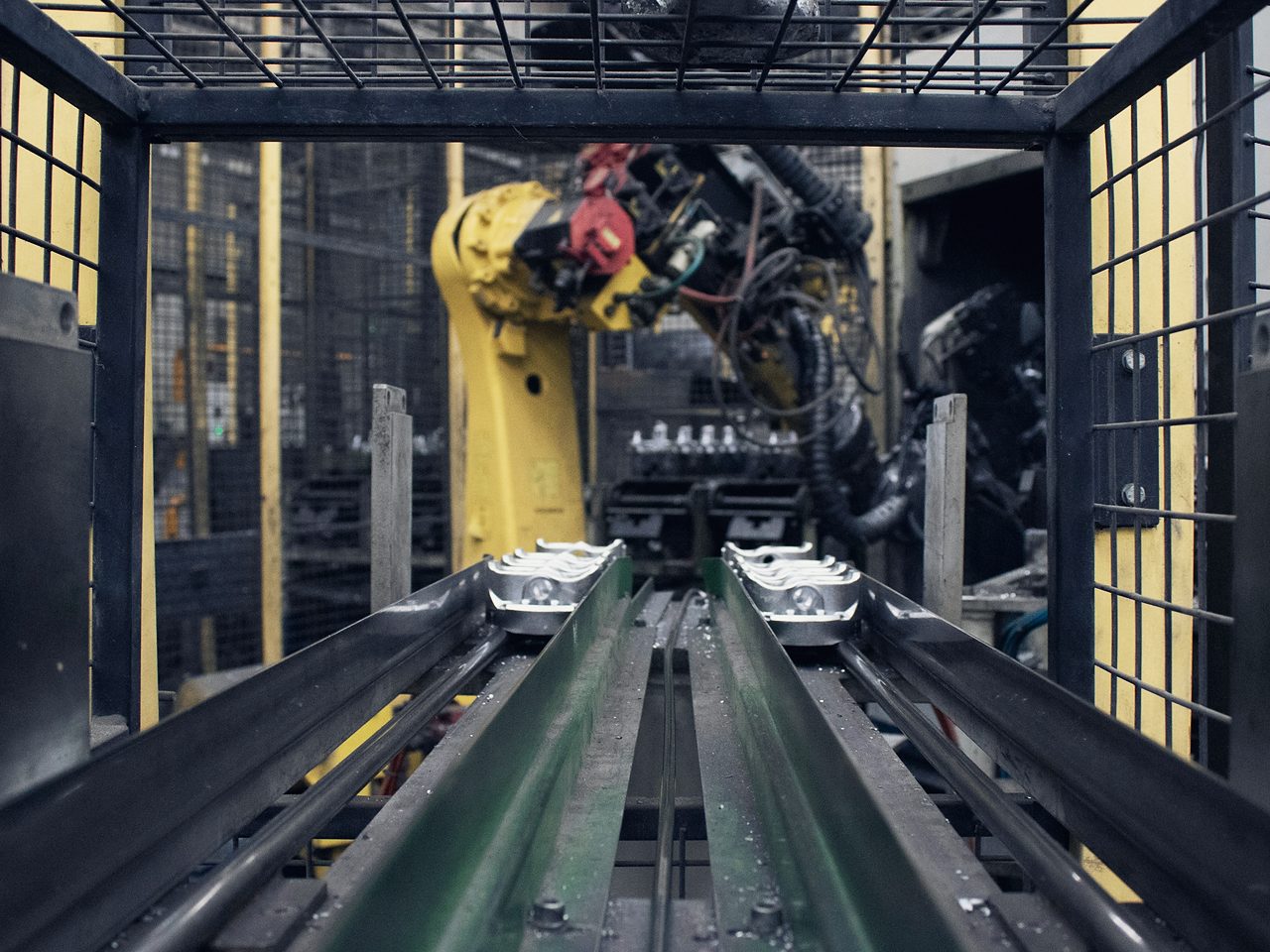 Celda robotizada con un brazo industrial amarillo; en primer plano, rieles que acercan piezas metálicas a la celda.